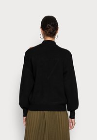 Zwarte gebreide trui met hoge kraag en ballonmouwen, met een geribbelde onderrand en manchetten. Draag het met een geplooide olijfgroene rok.