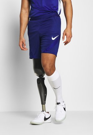 Homme portant un short et un t-shirt de sport bleus, chaussette blanche et basket, avec une jambe prothétique noire, faisant un pas en avant sur un fond uni.