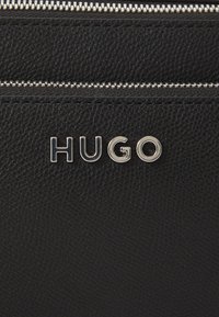 Juodas odinis krepšys su tekstūruotu paviršiumi ir sidabriniu užtrauktuku. Prekės ženklas "HUGO" ryškiai išdėstytas blizgiais metaliniais rašmenimis.