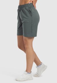 Pantalones cortos verde oscuro con una cinturilla elástica, que presentan dos bolsillos laterales y una tela texturizada. Combinados con zapatillas deportivas blancas.