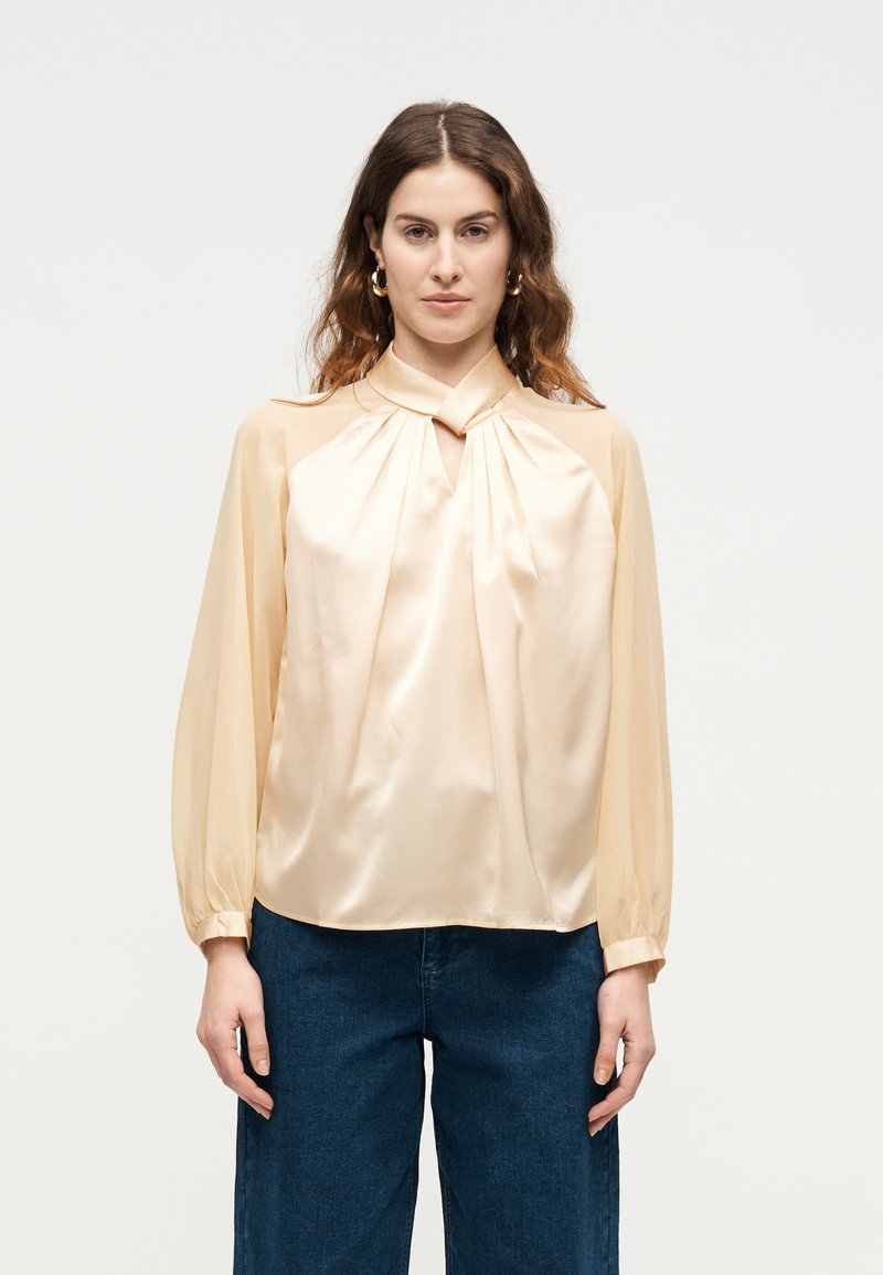 Γυναίκα που φοράει μπλούζα από μπεζ σατέν με μακριά μανίκια και άνοιγμα στο λαιμό, καθώς και σκούρο μπλε τζιν παντελόνι με ψηλή μέση, στέκεται μπροστά σε ένα απλό ανοιχτόφωτο φόντο.