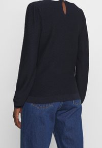 Marinfärgad långärmad topp i texturerat tyg, rund halsringning och liten nyckelhålssnitt bak. Matchad med blå jeans.