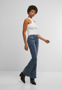 Jeans a zampa d'elefante blu scuro con vita media, tasche standard e dettagli di cucitura sottili. Abbinati a un top aderente bianco senza maniche e stivali neri alla caviglia.