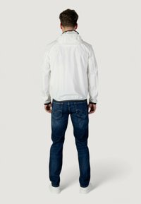 Giacca bianca impermeabile con cappuccio, polsini elasticizzati e cordino. Indossata con jeans blu scuro e scarpe sportive bianche. Vista da dietro.