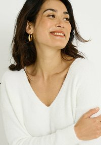 Pull blanc moelleux à col en V avec manches longues. Le modèle porte des boucles d'oreilles dorées en forme de créoles et a des cheveux foncés et ondulés. Fond neutre.