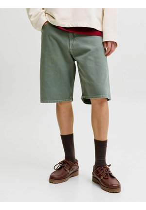 Muž nosiaci zelené šortky po kolená, tmavé ponožky, hnedé kožené topánky a krémovú košeľu s dlhým rukávom a červeným tričkom pod ňou.