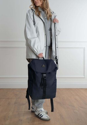 Navyblå rygsæk med fold-over låg, justerbare stropper og et lille logo. Bæres med en hvid regnjakke og grå sweats.
