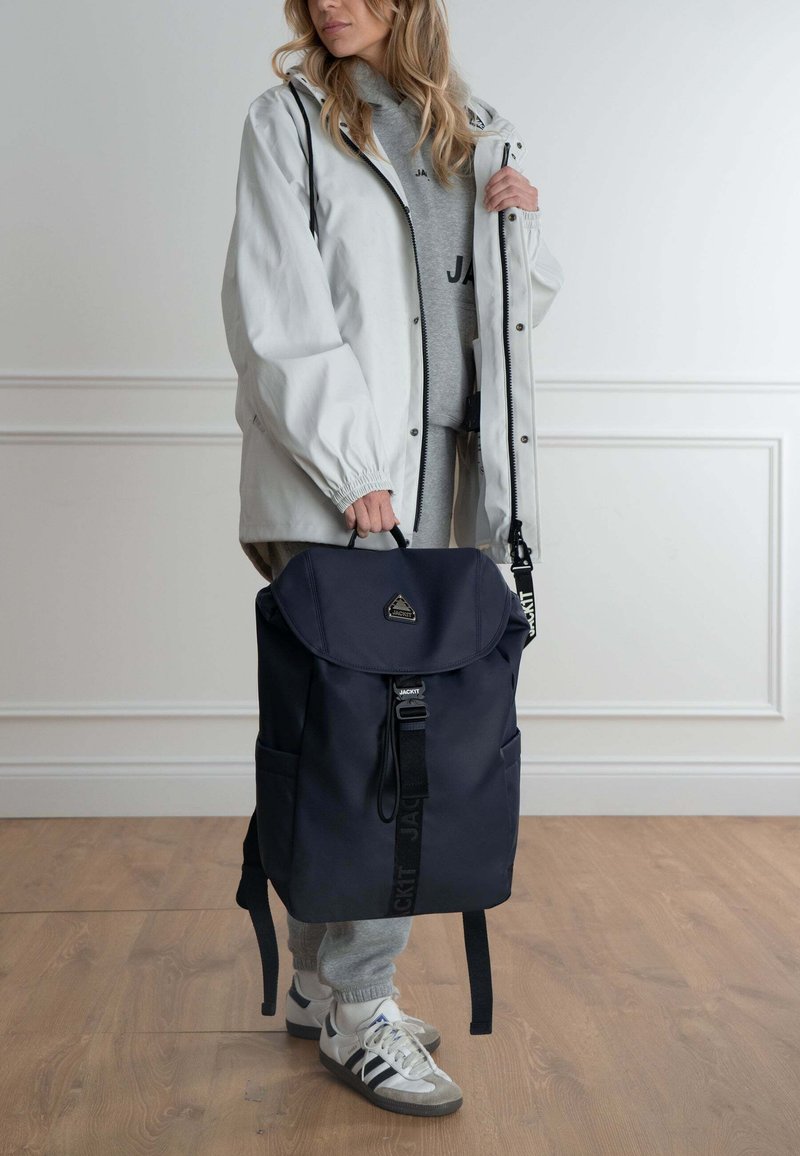 Zaino blu navy con patta pieghevole, spallacci regolabili e un piccolo logo. Indossato con un impermeabile bianco e pantaloni della tuta grigi.