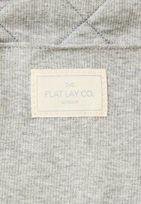 Grå ribbet stoff med en quiltet tekstur har en kremfarget vevet etikett merket "THE FLAT LAY CO. LONDON" sydd på den ene siden.