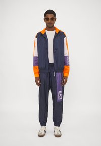 Herre treningsdrakt i marineblå med oransje, lilla og hvite detaljer. Har en zipjakke og bukser med snor i livet, laget av lettvevde materialer. Sneakers inkludert.