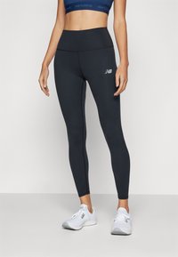 Leggings taille haute noirs en matériau extensible avec une légère brillance, dotés d'un petit logo blanc sur la cuisse. Associés à des chaussures blanches.