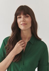 Cămașă verde cu guler, cu nasturi, textură moale, croială lejeră. Modelul are părul lung și ondulat și ține gulerul cămășii cu o mână.