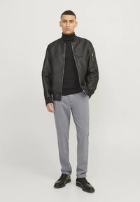 Schwarze Leder-Bomberjacke über einem schwarzen Rollkragenpullover, kombiniert mit hellgrauen Hosen und schwarzen Schuhen. Merkmale sind Reißverschlüsse und ein tailliertes Design.