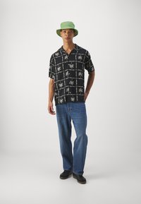 Camisa negra de manga corta con patrón de hojas de palma blancas, combinada con jeans azul claro y un sombrero de cubo verde. Zapatos negros completan el conjunto.