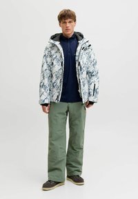 Veste isolante pour homme avec un motif marbré blanc et bleu, des accents noirs, associée à un pantalon imperméable vert et des chaussures sombres.