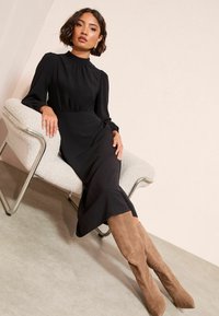 Melna garām piedurknēm tērps ar augstu apkakli, savāktu jostasvietu un plūstošu svārku, apvienots ar brūnu zamša augstiem zābakem, sēžot krēslā.