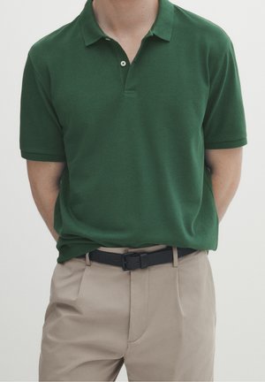 Grünes Poloshirt aus weichem Stoff mit einem Zwei-Knopf-Verschluss und kurzen Ärmeln, kombiniert mit beigen Hosen und einem schwarzen Gürtel.
