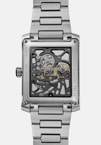 Srebrny zegarek ze stali nierdzewnej z prostokątną kopertą, przezroczystym szkieletem tarczy oraz widocznymi zębatkami, z metalową bransoletą i wodoodpornością.