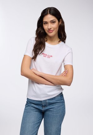 T-shirt bianca a maniche corte, colletto rotondo, con testo ricamato rosso "ANSIA CLUB H24/7". Abbinata a jeans di denim azzurro chiaro.