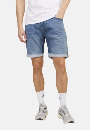 Hombre con pantalones cortos de mezclilla azul remangados, calcetines blancos hasta el tobillo, zapatillas grises y camiseta blanca, con una mano en el bolsillo.