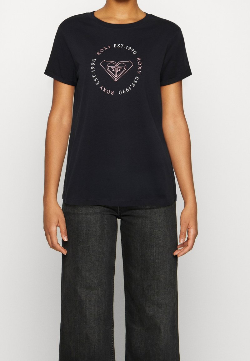 Personne portant un T-shirt noir Roxy avec un logo circulaire rose et le texte "ROXY EST. 1990", associé à un jean large foncé.