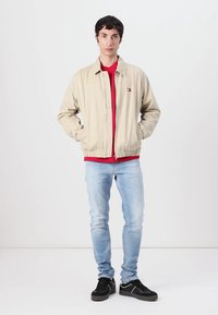 Beige bomberjakke med glidelås foran, rød lag under, lyseblå jeans og svarte sneakers. Enkel design med avslappet passform.