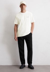 White cotton t-shirt with subtle text print, black linen pants, and dark shoes, complemented by a light-colored baseball cap.