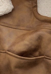 Ruskea keinonahkainen materiaali, jossa on pehmeä, vaaleanvalkoinen fleece-kaulus. Sisältää näkyvää ommelta ja taskuyksityiskohtia edessä.