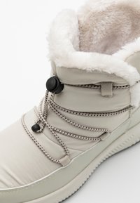 Beige vinterstøvle med kunstpels foring, elastiske snørebånd og mønstret sål. Har en snorelukning og polstret ankel for komfort.