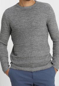 Pull en maille grise texturée avec motifs à côtes horizontales, col rond et poignets ajustés, porté avec un pantalon bleu. Finition en tissu doux.