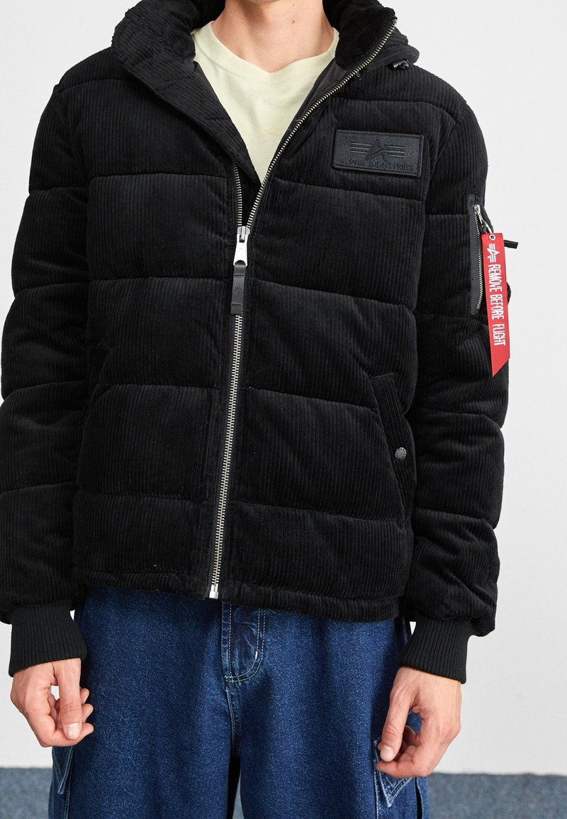 Černá hedvábná puffer bunda s prošívaným designem, zipem vpředu, kapucí a výraznou nášivkou loga. Na boční kapse je červený štítek.