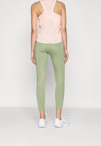 Débardeur rose clair avec un dos nageur associé à un legging vert comportant des panneaux en maille et des accents cousus, porté avec des chaussures de sport blanches.