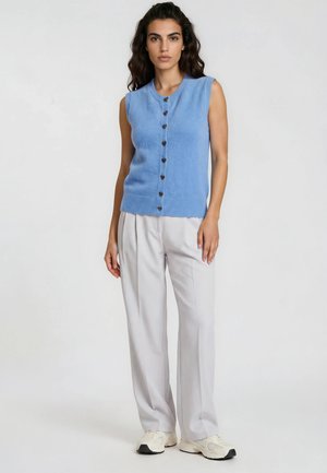 Woman with dark hair wearing a sleeveless blue buttoned sweater, light gray wide-leg pants, and white sneakers standing against a white background.