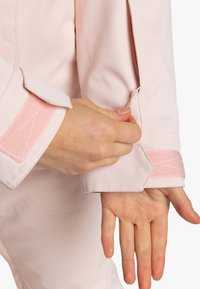 Lichtroze stoffen blouse met een ritsmanchet, versierd met een roze klittenbandaccent en een hexagonaal tabdetail op de mouw.