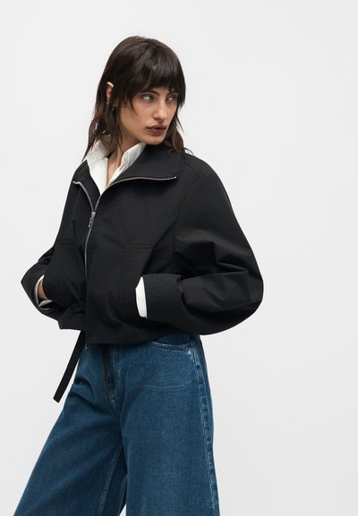 Γυναίκα με σκούρα μαλλιά φοράει μαύρο κοντό μπουφάν πάνω από λευκό πουκάμισο και φαρδιά μπλε τζιν, στέκεται με τα χέρια στις τσέπες, κοιτάζοντας αριστερά.