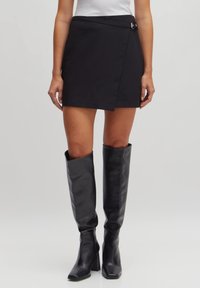 Zwarte wikkelrok gemaakt van gladde stof met een zijsluiting en een strakke silhouet, gecombineerd met zwarte leren laarzen tot de knie.