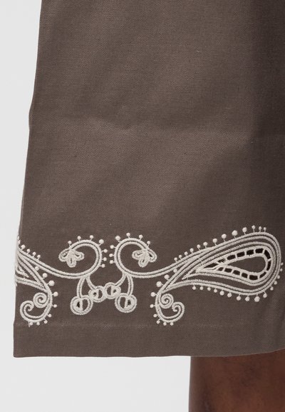 Καφέ ύφασμα με λευκή περίτεχνη κεντητική που περιλαμβάνει σπείρες, θηλιές και διακοσμητικές βούλες κατά μήκος της ακμής του υφάσματος.