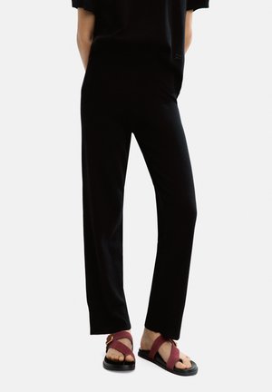 Czarne spodnie w szerokim kroju o gładkiej fakturze i elastycznym pasie, zestawione z bordowymi sandałami z paskiem i złotymi elementami.