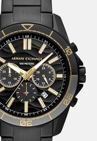 Črni kronografski ure s zlatimi poudarki in teksturiranim črnim številčnico. Ima tri podštevce, prikaz datuma in vodoodpornost do 100 metrov.