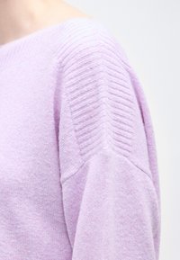 Κοντινό πλάνο ενός ατόμου που φοράει ένα απαλό λιλά πουλόβερ με ριμπ λεπτομέρειες στους ώμους σε ανοιχτό φόντο.