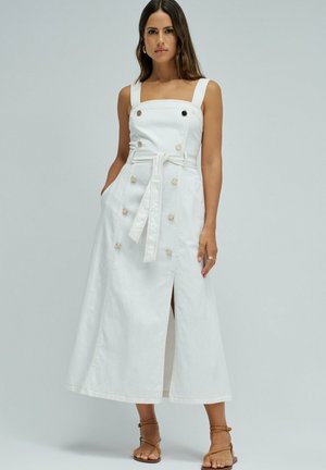 Mujer con vestido midi blanco sin mangas abotonado, con tirantes anchos y cinturón anudado en la cintura, de pie con las manos en los bolsillos.