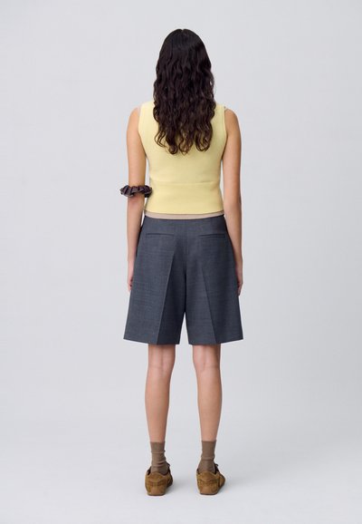 Top giallo a maniche corte lavorato a maglia e pantaloni corti grigi a pieghe visti da dietro. I dettagli includono un elastico marrone sul braccio e scarpe marroni con calze beige.
