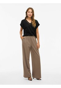 Frau steht mit einer Hand in der Tasche, trägt ein schwarzes kurzärmeliges Oberteil und weite, taubfarbene Hosen mit schwarzen hohen Sandalen.