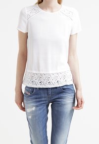 Top corto bianco a maniche corte con orlo in pizzo floreale e dettagli tagliati al laser sulle spalle, abbinato a jeans blu strappati.
