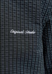 Μαύρο ύφασμα με λευκό σχέδιο πλέγματος και λευκή κεντημένη επιγραφή που αναγράφει "Originals Studio" στην περιοχή του στήθους.