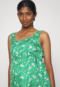 Vestido floral verde com silhueta ajustada, decote arredondado e detalhe de laço lateral. O tecido tem uma textura suave e padrões vibrantes.