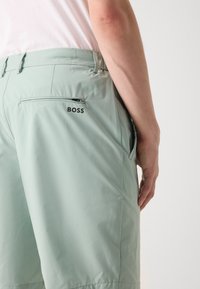 Lysegrønne shorts med en glat tekstur, der har en lynlås baglomme og et trykt "BOSS" logo nær taljen.