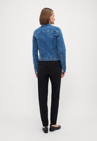 Lys blå denimjakke med en tettsittende design, klassisk krage og synlige sømdetaljer, kombinert med sorte bukser og flate sko.