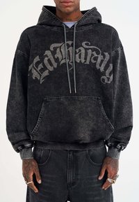 Black hooded sweatshirt in distressed denim with grey lettering. Features a front pocket and ribbed cuffs. Casual, oversized fit.