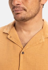Short-sleeve, button-up shirt in light orange, featuring a soft, textured fabric, a collar, and a single button detail.